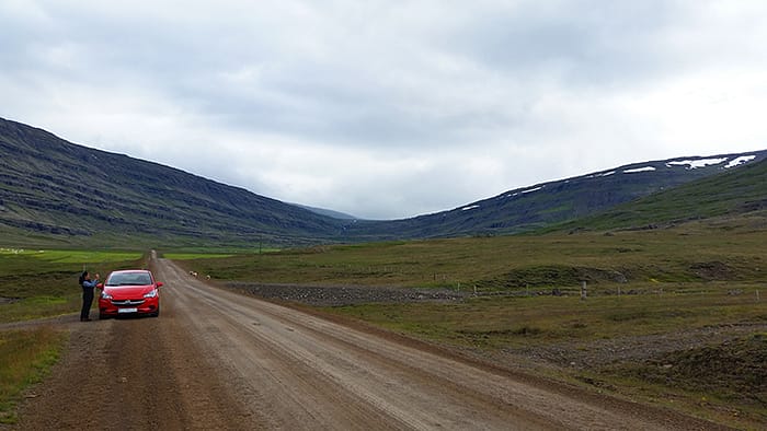 image of a red car on a dirt road with green hills in the background in Iceland. Planning a European road trip is the only way to see places like this.